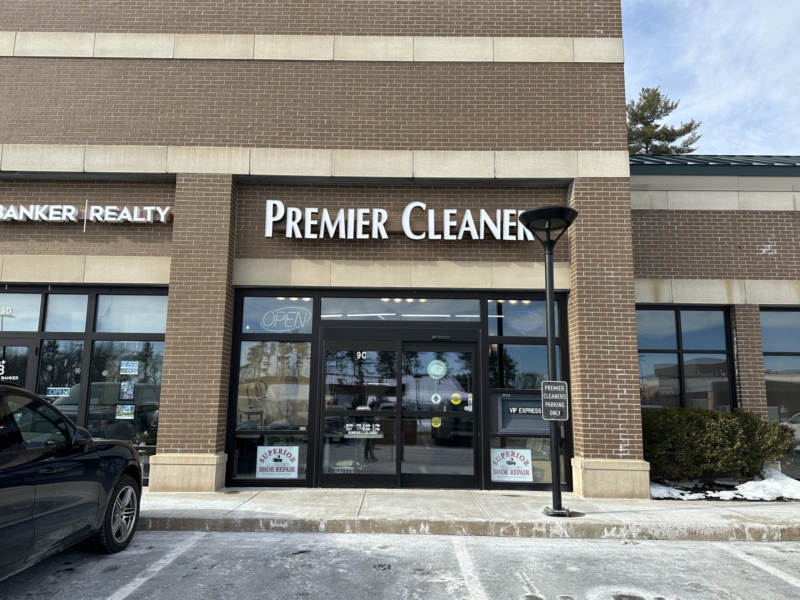 Storefront of Premier Cleaners with glass doors and windows. The sign is prominently displayed above. A few cars are parked nearby, and a sign indicates reserved parking for dry cleaning customers.