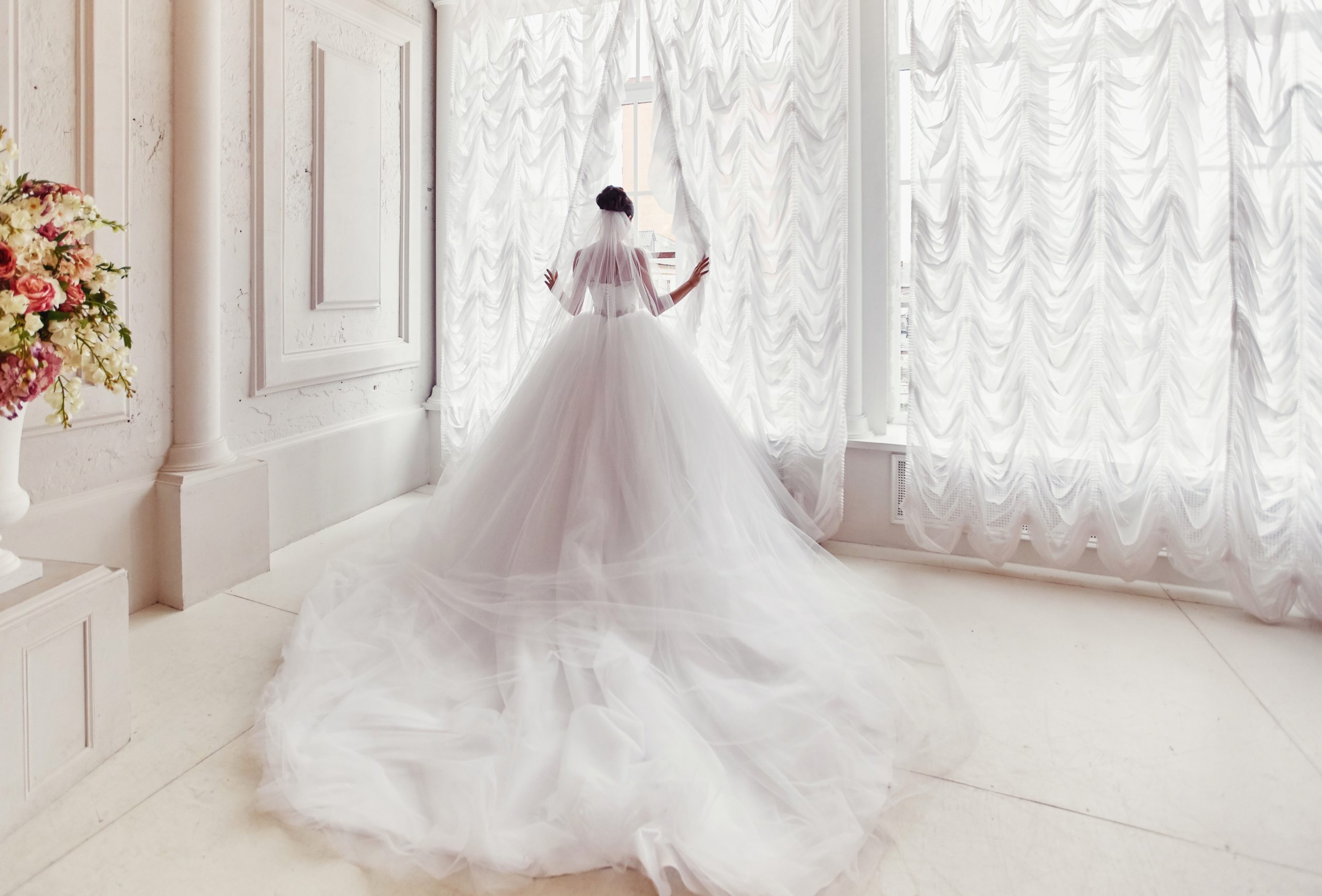 A bride in a voluminous white gown stands facing sheer curtains, holding them slightly open.