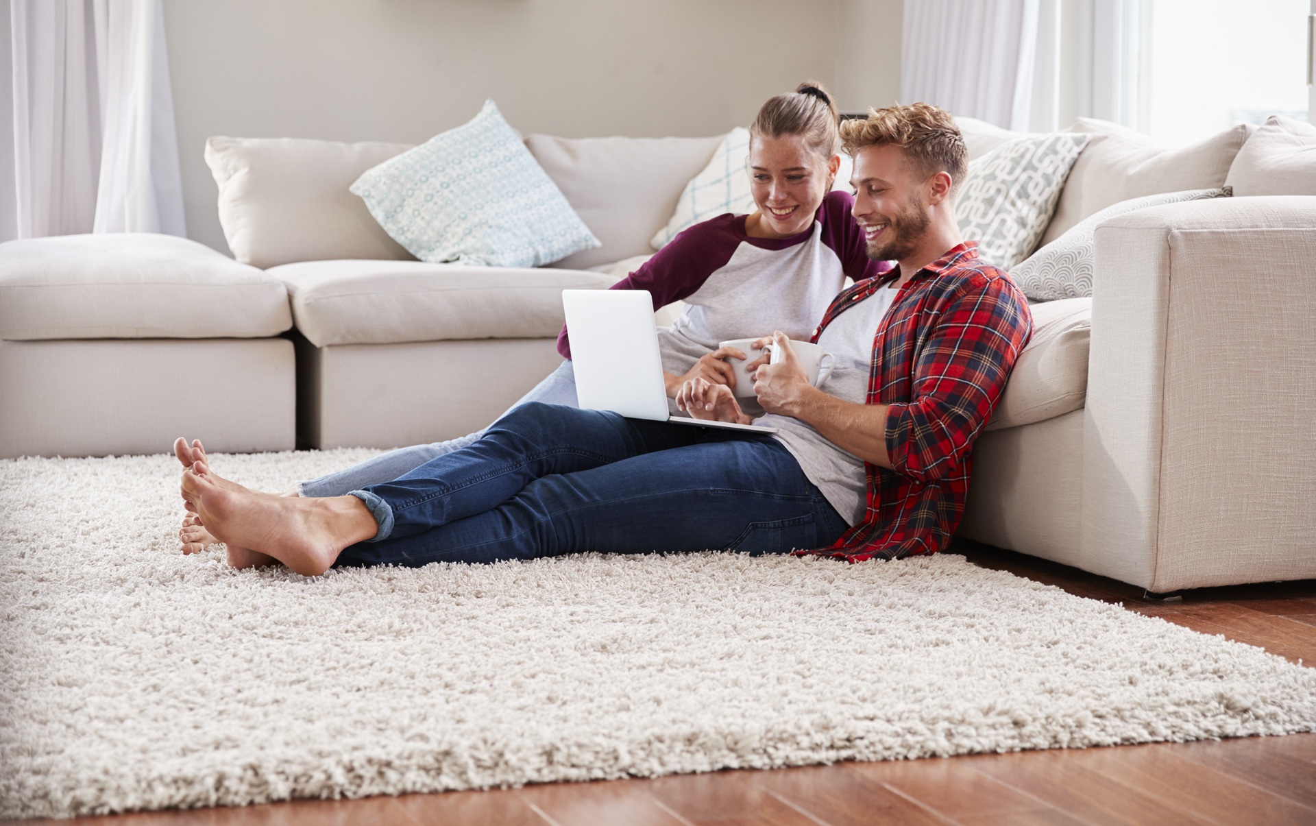 A couple sits on the floor, leaning against a couch, using a laptop. They are smiling and appear relaxed in a cozy living room setting.