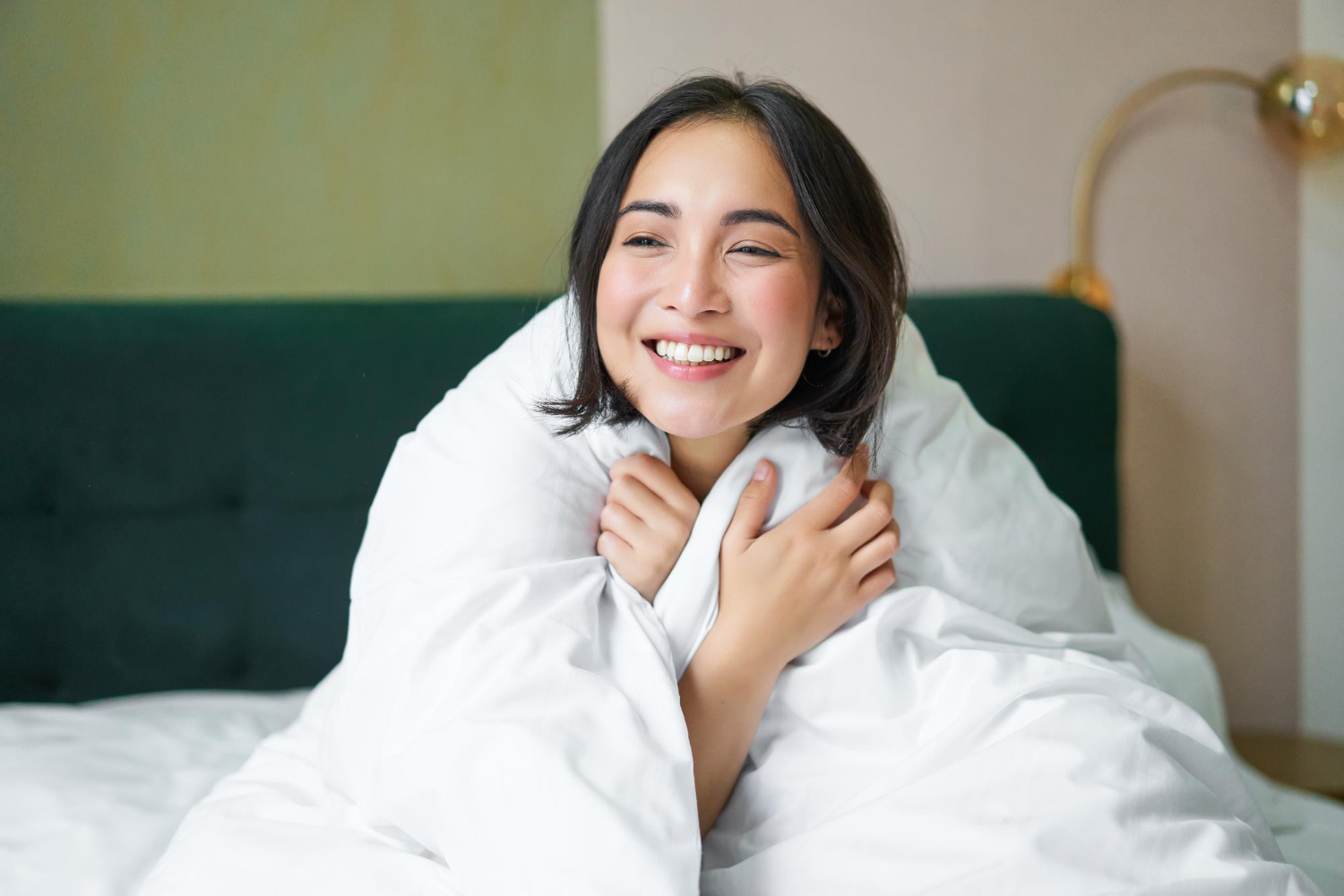 Person smiling while wrapped in a white blanket, sitting on a bed. A lamp is visible in the background.