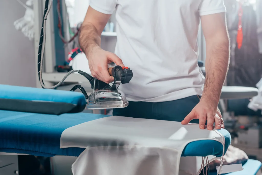 A person in a white t-shirt is ironing a piece of fabric on a blue ironing board in a well-lit room, showcasing the care you'd expect from a top laundry service near me.