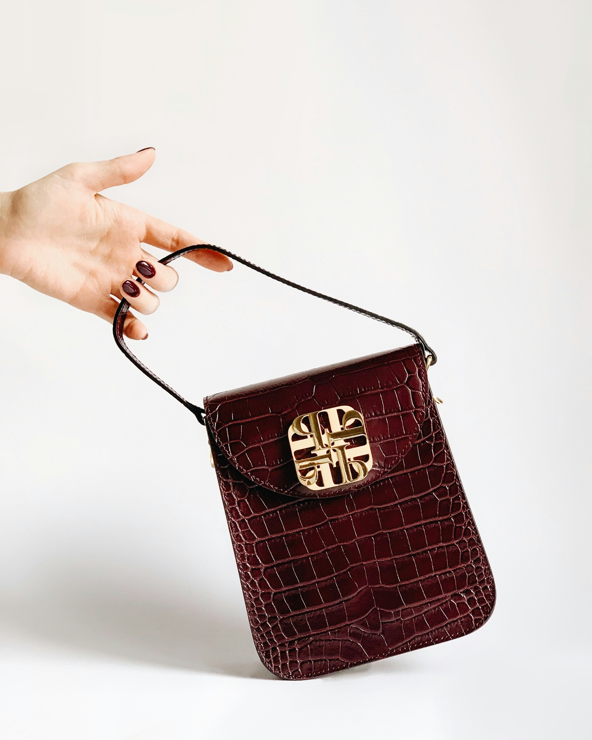 A hand holds a burgundy crocodile-patterned handbag with a gold decorative clasp against a white background.
