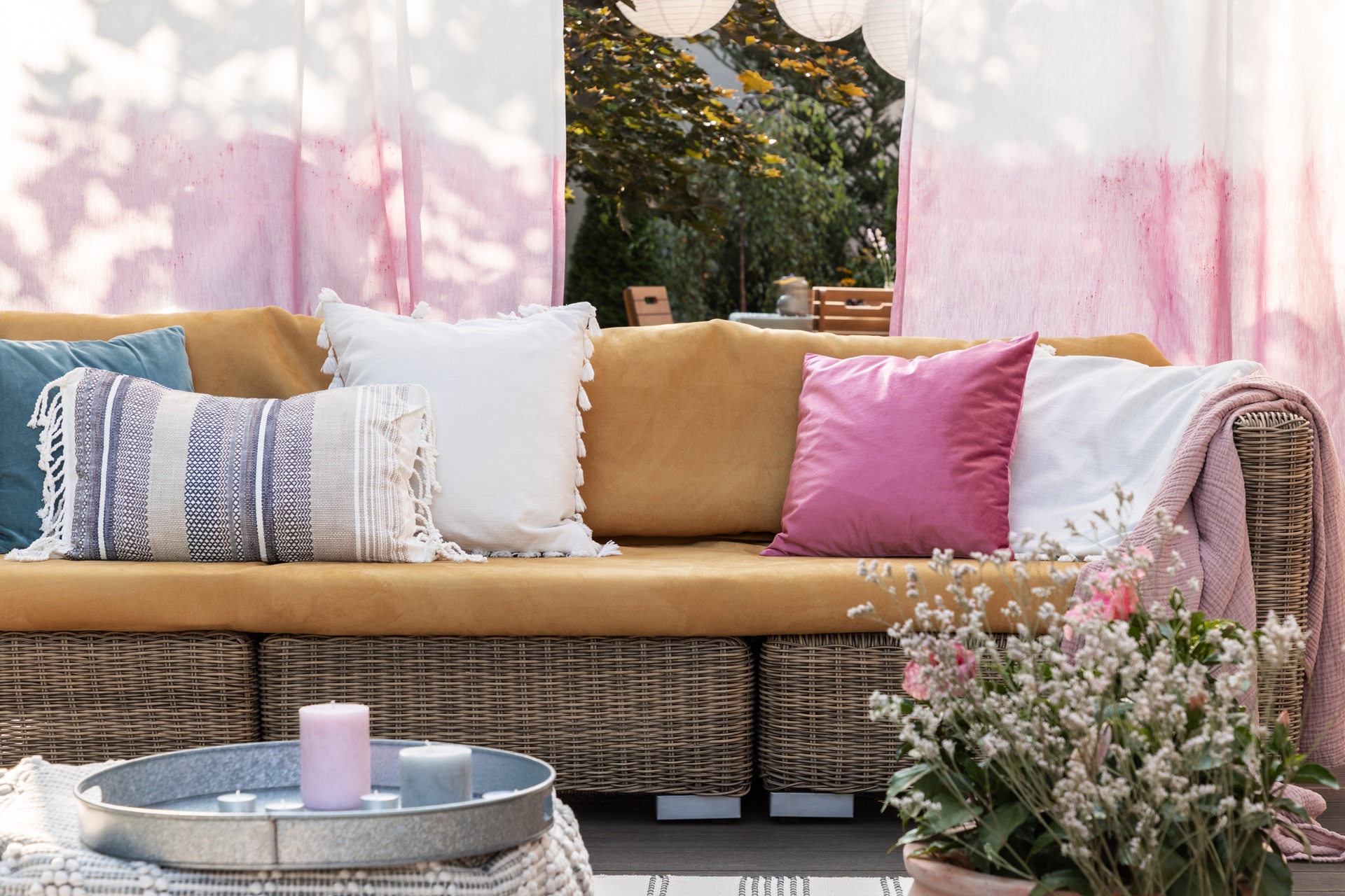 Outdoor wicker sofa with yellow cushions, adorned with white, pink, blue, and striped throw pillows. Nearby, a metal tray with candles and a plant in the foreground. Pink curtains hang behind.