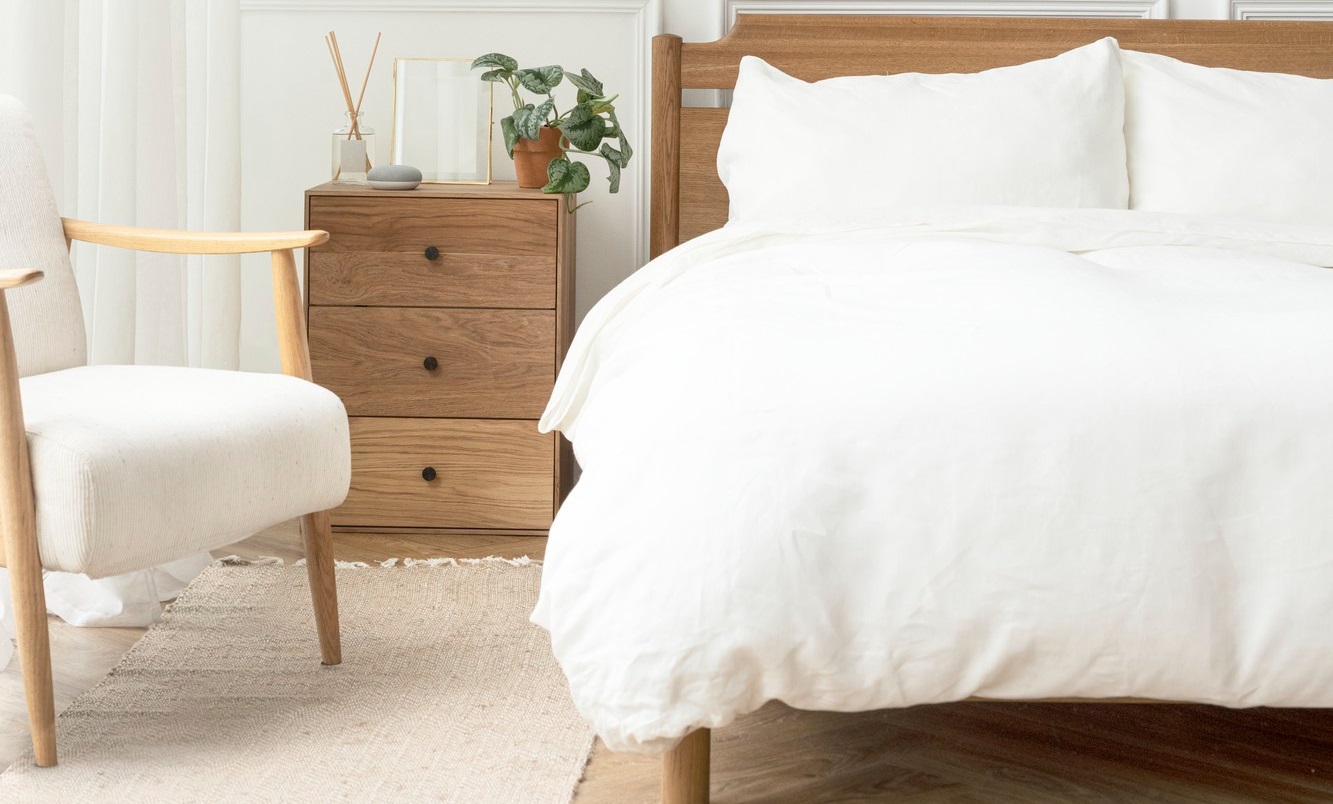 A neatly made bed with white bedding is next to a wooden bedside table with a plant and decor. A wooden chair with a cushioned seat is on a beige rug.