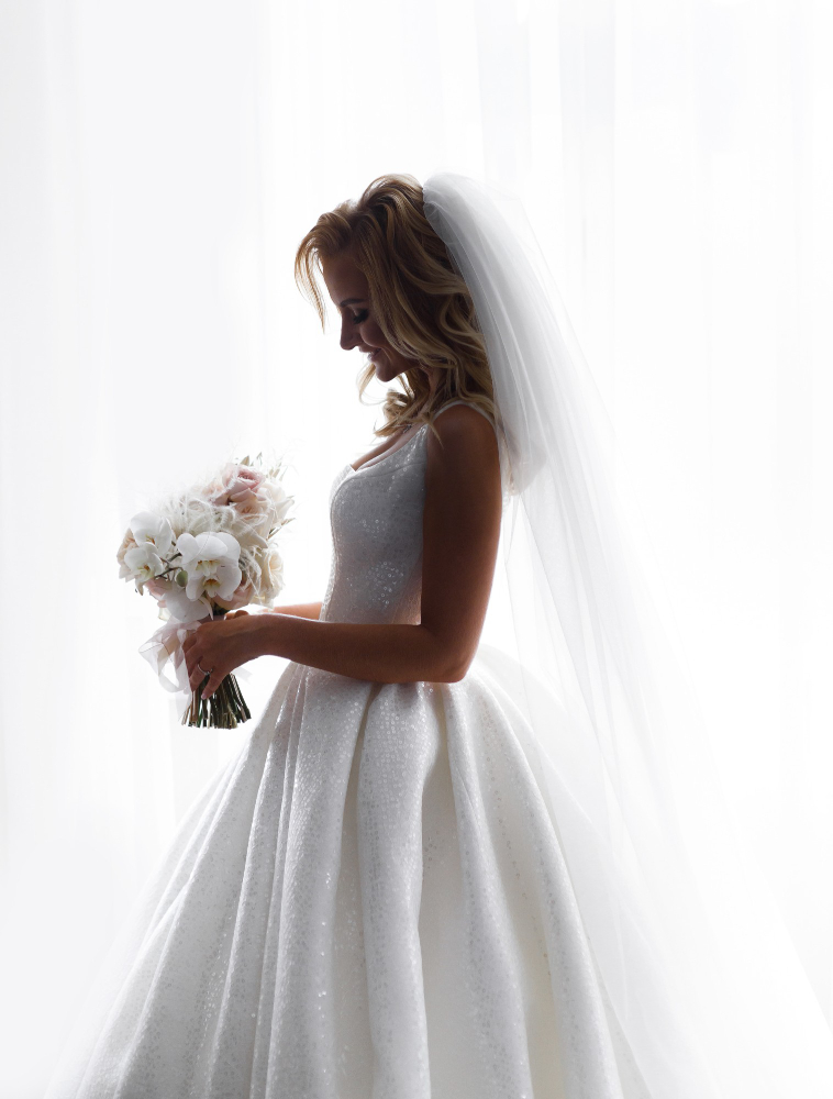 Bride in a white gown and veil holds a bouquet of flowers, standing in front of a bright window.