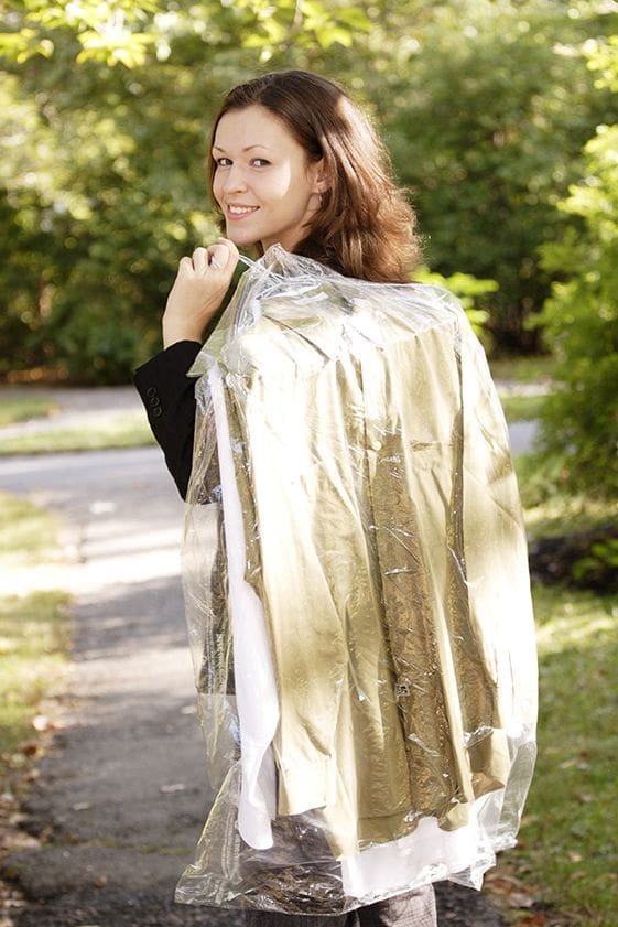 A person outdoors holding a suit covered in dry cleaning plastic over their shoulder, turning to look back and smiling. Trees and a path are in the background.