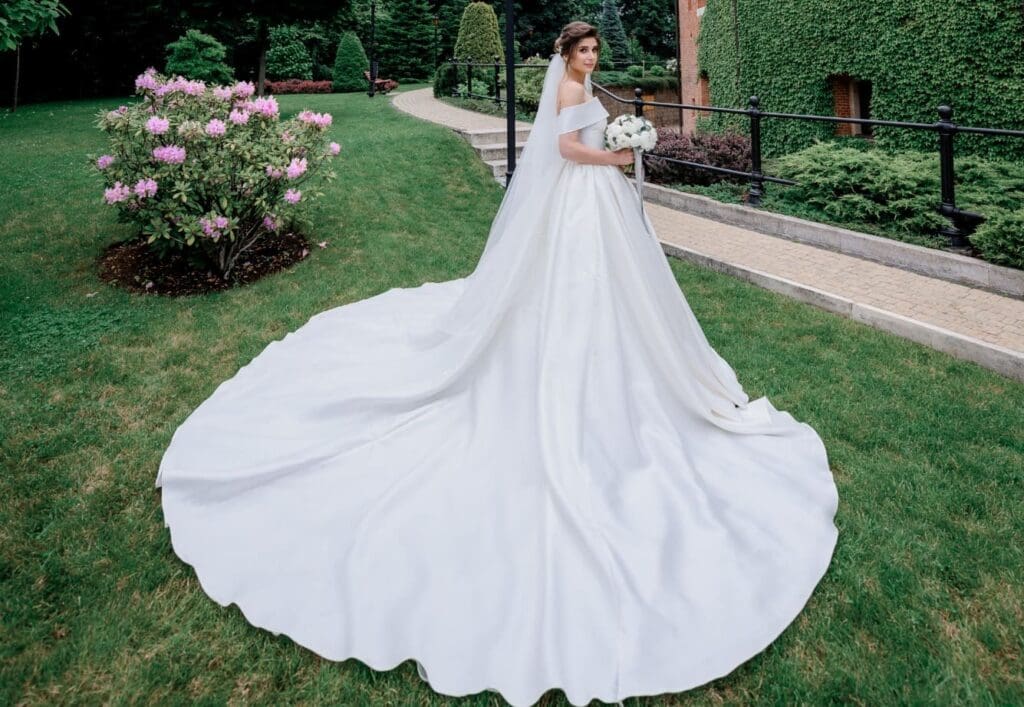 A bride in a white dress with a long train stands on a grassy lawn, holding a bouquet, with flowers and greenery in the background.