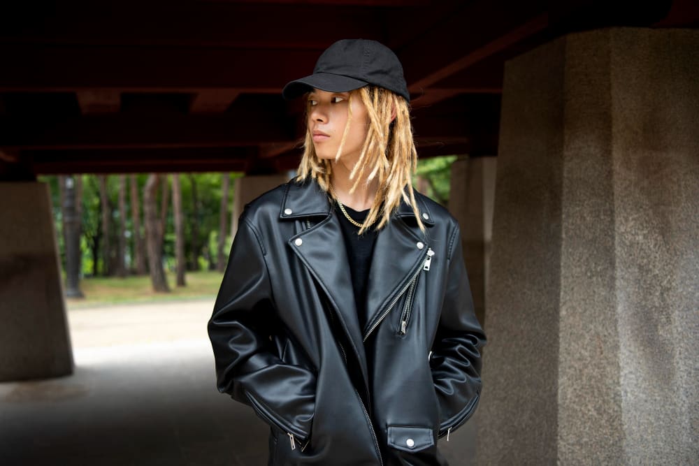 Person with blond dreadlocks wearing a black cap and black leather jacket stands outdoors under a structure, looking to the side. Trees are visible in the background.