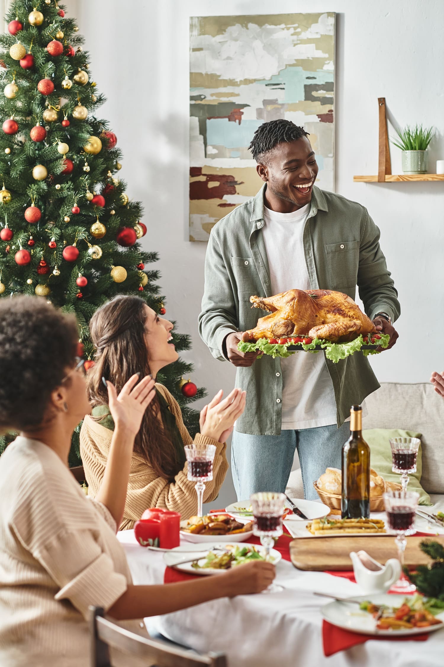 A man carries a roast turkey to a festive dining table with three people seated; a decorated Christmas tree is in the background.