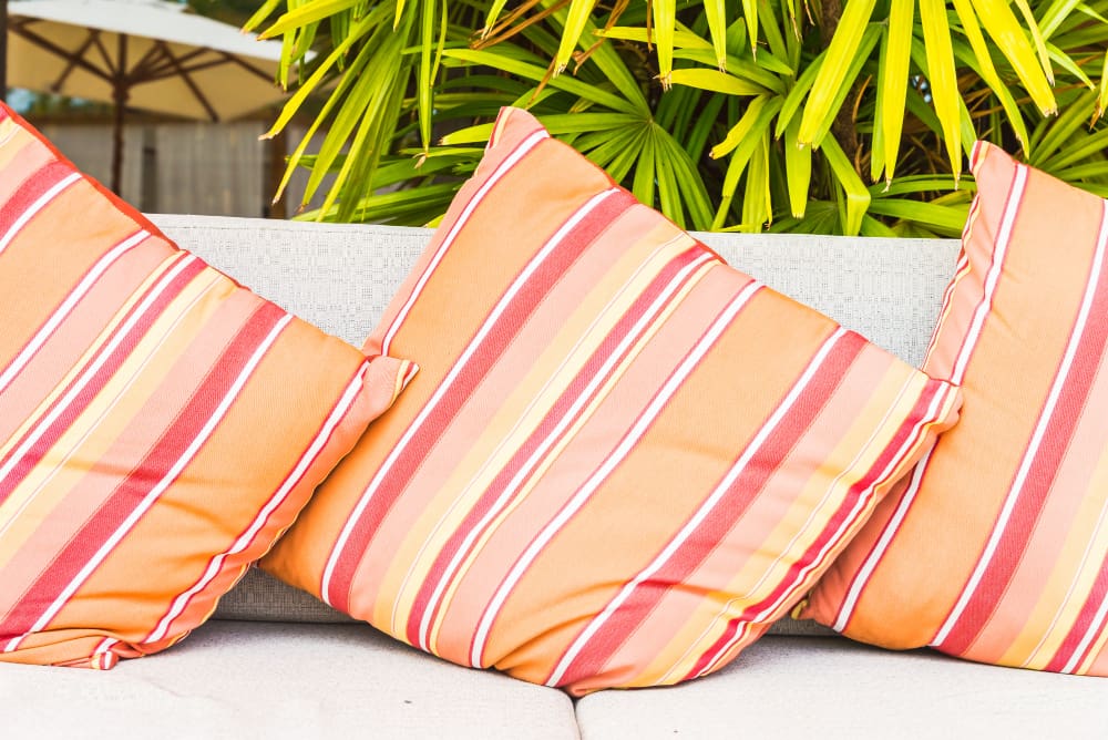 Three striped orange, yellow, and red pillows are arranged on a light-colored outdoor sofa, with green leafy plants and an umbrella in the background.