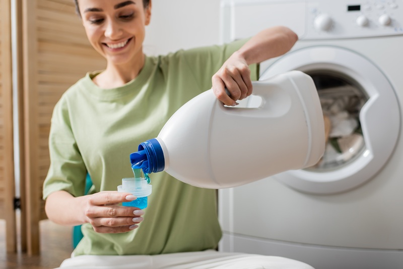 A person pours blue liquid laundry detergent from a large bottle into a measuring cap in front of a washing machine.
