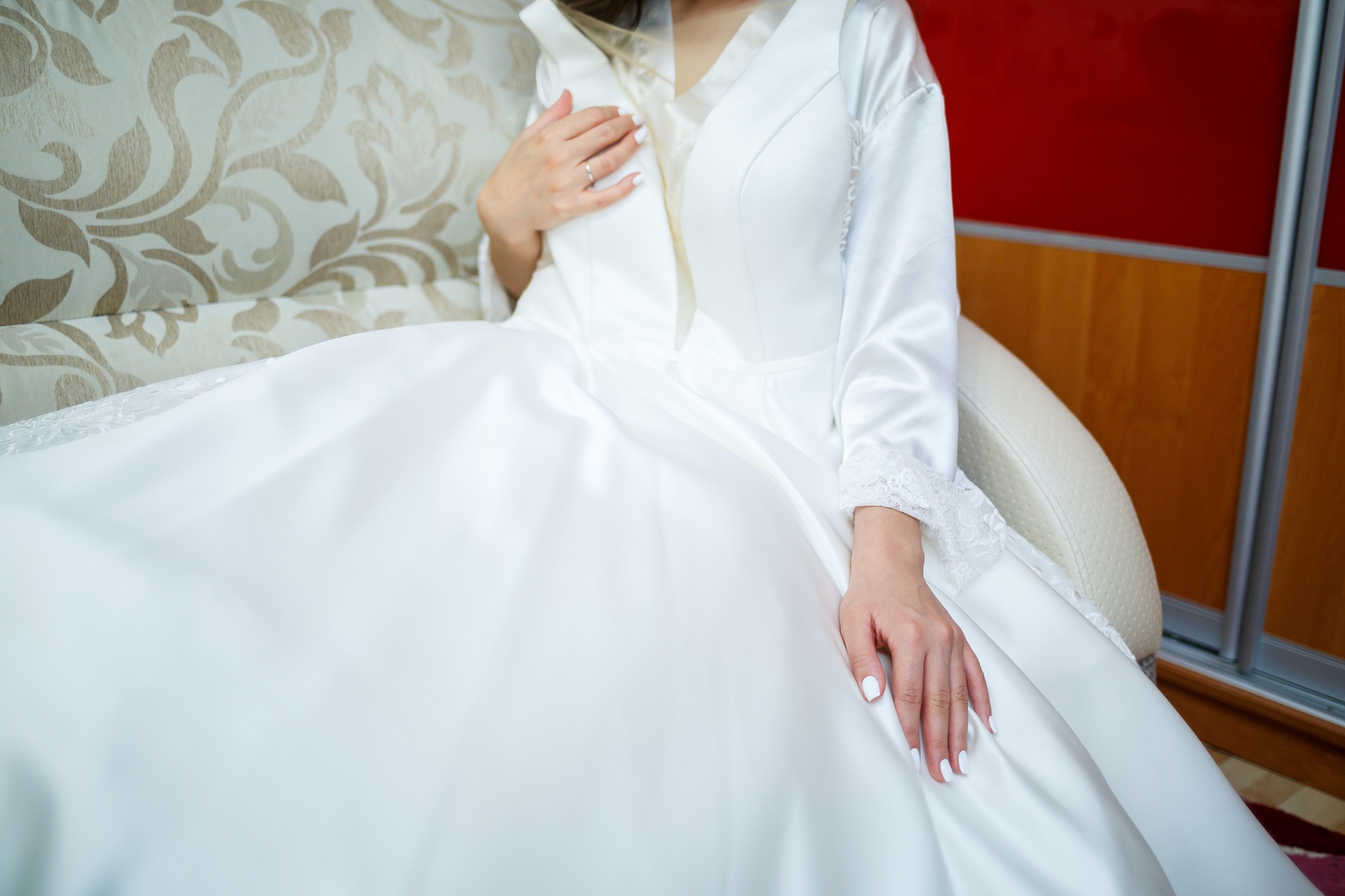 A person in a white wedding dress sits on a patterned sofa with hands resting on their lap, showing manicured nails.