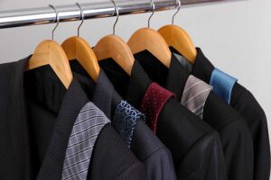 Five suits on hangers, each paired with a different patterned or colored tie, hanging on a metal clothing rack against a plain background.