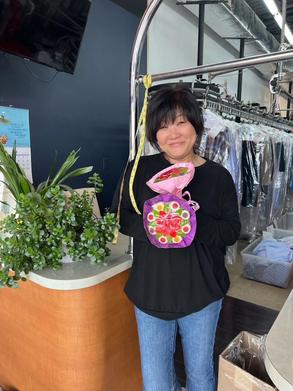 A woman stands in a dry cleaning shop, holding a colorful flower bouquet in a pink bag, with clothes hanging on racks and a plant on the counter beside her.