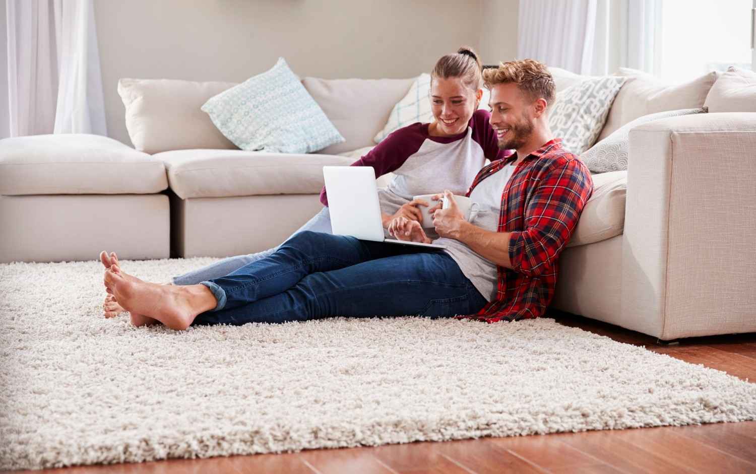 Two people sit on a carpeted floor, leaning against a sofa, looking at a laptop and smiling in a bright living room.