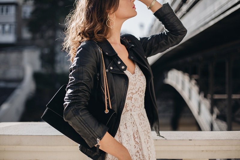 Woman wearing a black leather jacket over a white lace dress stands near an outdoor railing, holding a black handbag, with buildings and a bridge in the background.