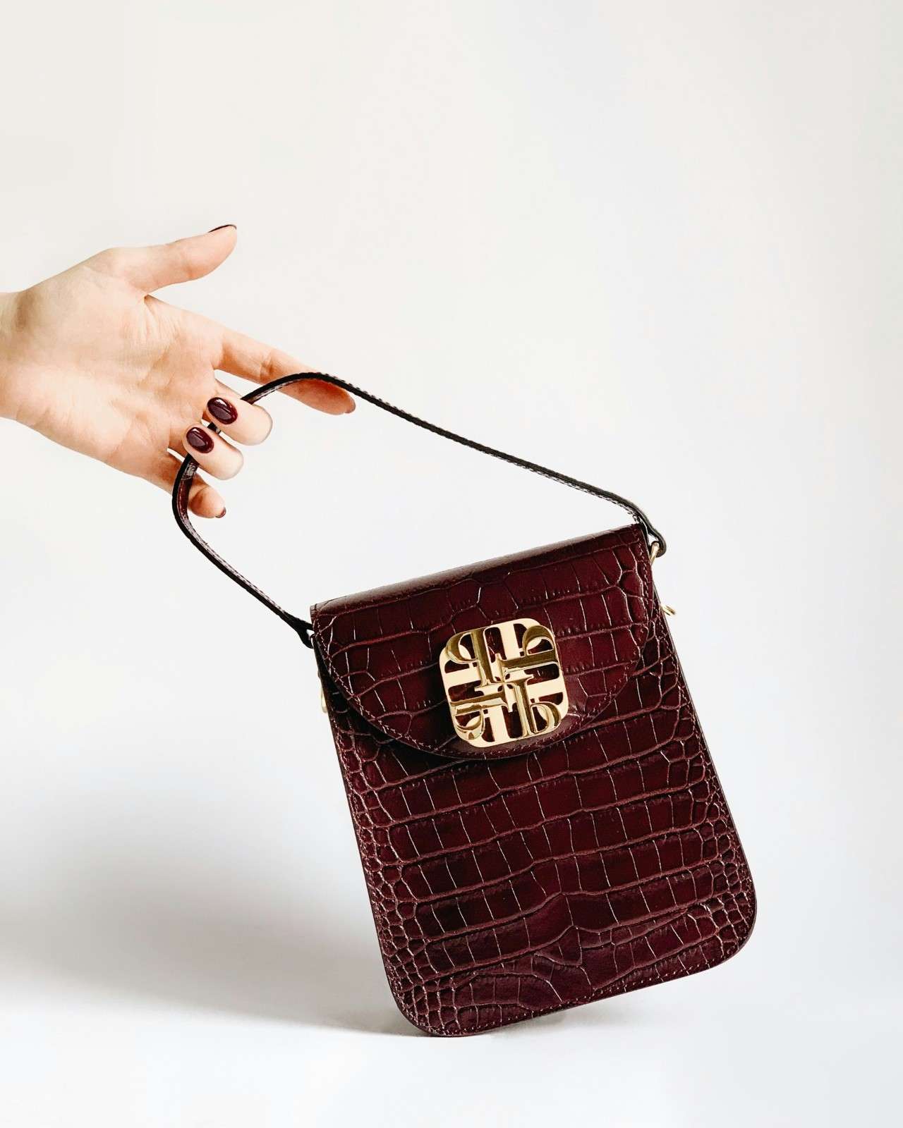 A hand holds a dark brown crocodile-patterned handbag with a short strap and a large gold decorative clasp against a plain white background.