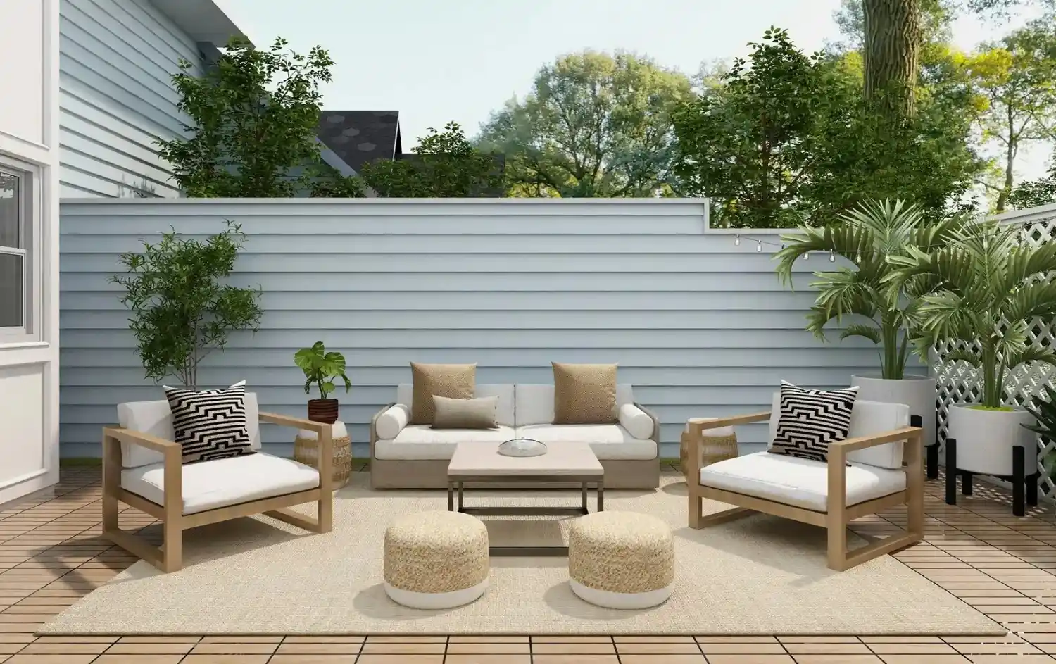 Outdoor patio with light wood furniture, beige cushions, two round ottomans, potted plants, and a white privacy fence in the background.