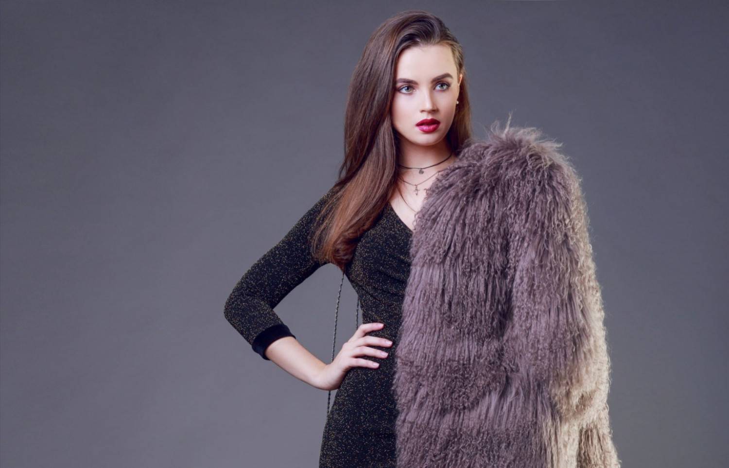 Woman with long brown hair wearing a dark dress and holding a large, shaggy gray coat, standing against a plain gray background.